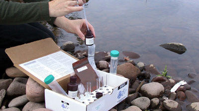 Person conducting water quality testing by a body of water with equipment and samples.